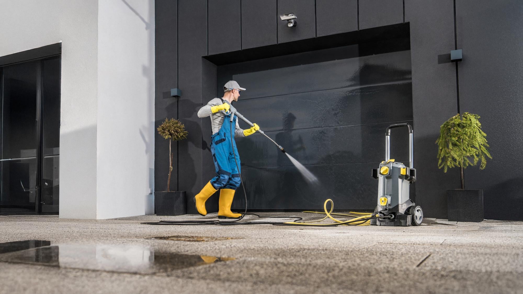 A person in blue overalls and yellow boots uses a pressure washer to clean a garage door, with a water hose connected to the machine. The setting features a modern building, and there are potted plants nearby.